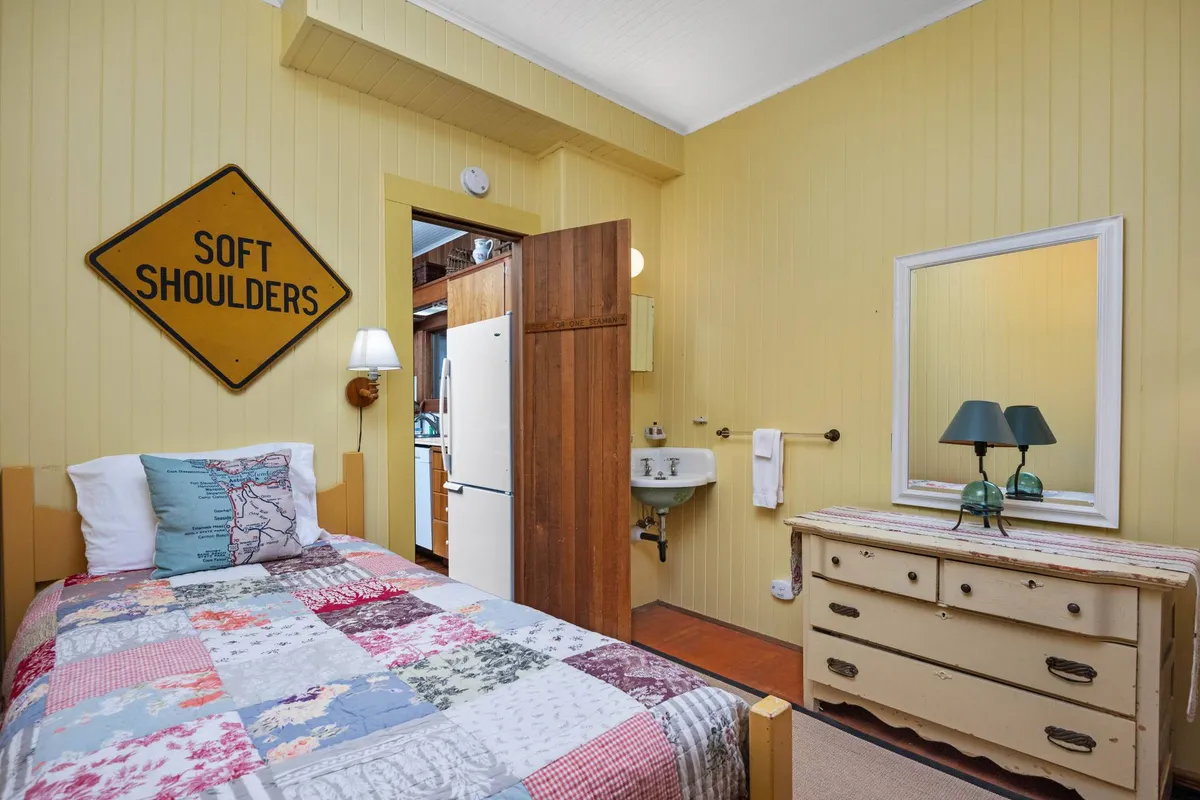 A cozy bedroom with a bed dressed in a quilted cover, a vintage dresser, and a nostalgic 'SOFT SHOULDERS' sign. The space feels comfortable, warm, and personal, with understated vintage touches that echo the retreat's calming decor.