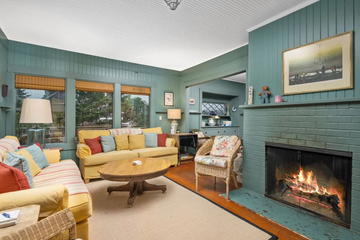 A cozy living room featuring a fireplace, a comfortable sofa with colorful cushions, a round wooden coffee table, and large windows letting in natural light. The space feels warm and inviting, suited for group gatherings and family game nights.