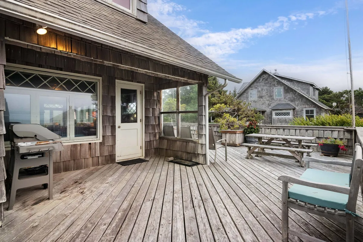 A welcoming deck area with outdoor seating and a grill, overlooking surrounding greenery. The setup evokes a casual, relaxed atmosphere ideal for outdoor dining, family barbecues, and unwinding after a day at the beach.