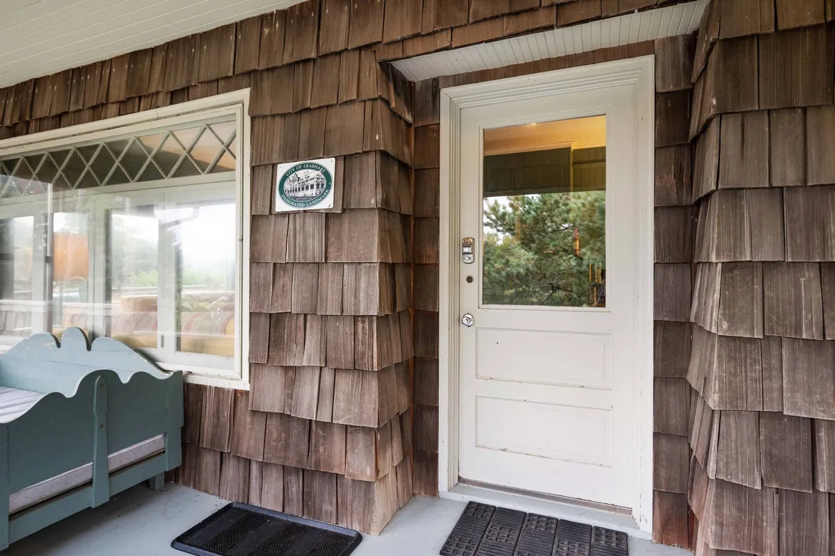 The front porch of Gin Ridge Cottage features a wooden shingle exterior, a welcoming front door, a decorative sign, and a cozy seating area. The nook is shaded and inviting, perfect for morning coffee, afternoon naps, or sharing quiet moments with loved ones (including furry friends).
