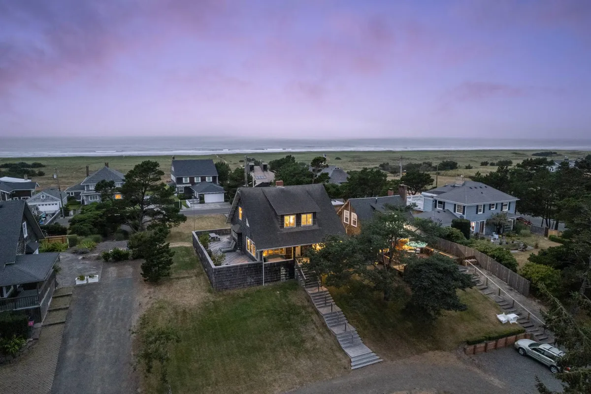 A broad view showing the cottage surrounded by lush trees with the Pacific Ocean visible in the distance. The vantage point captures both the natural beauty of the setting and the home's connection to the coastline.