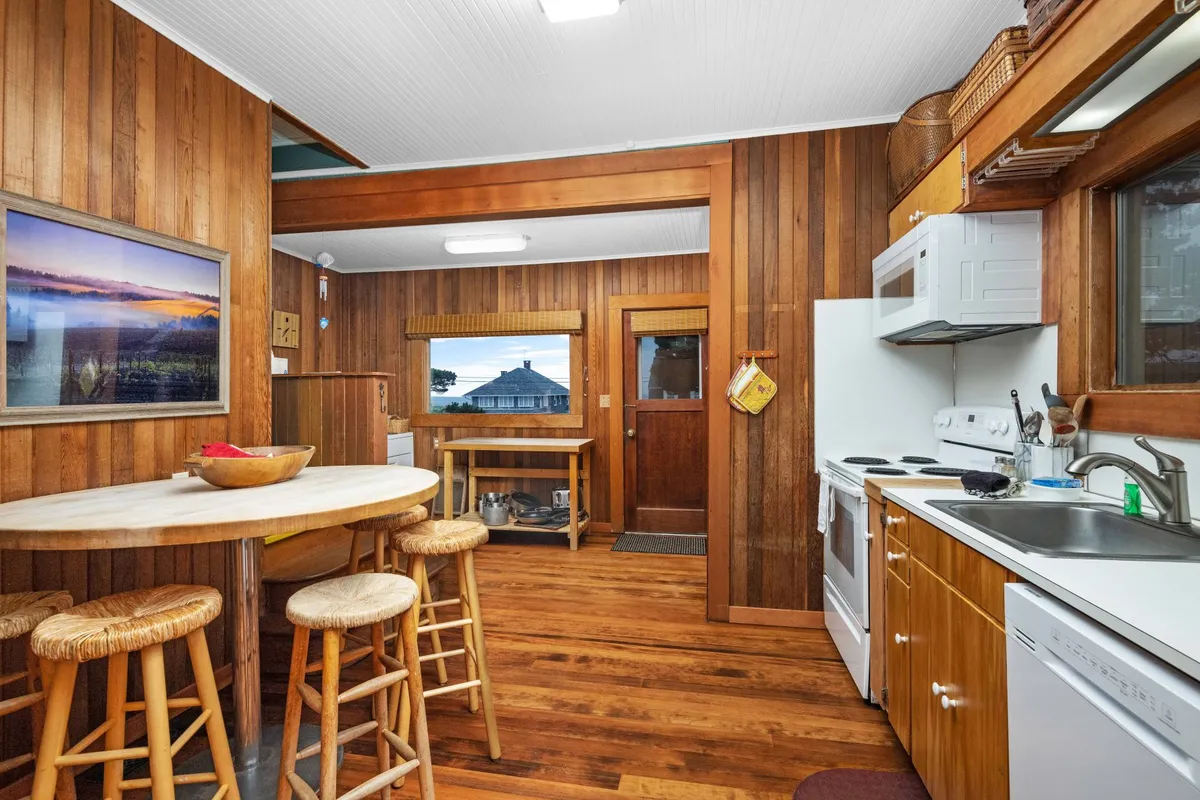A bright, inviting kitchen scene featuring a wooden round table with bar stools, modern kitchen appliances, and a framed landscape photograph. The space feels vibrant and ready for shared meals, casual breakfasts, or family celebrations.