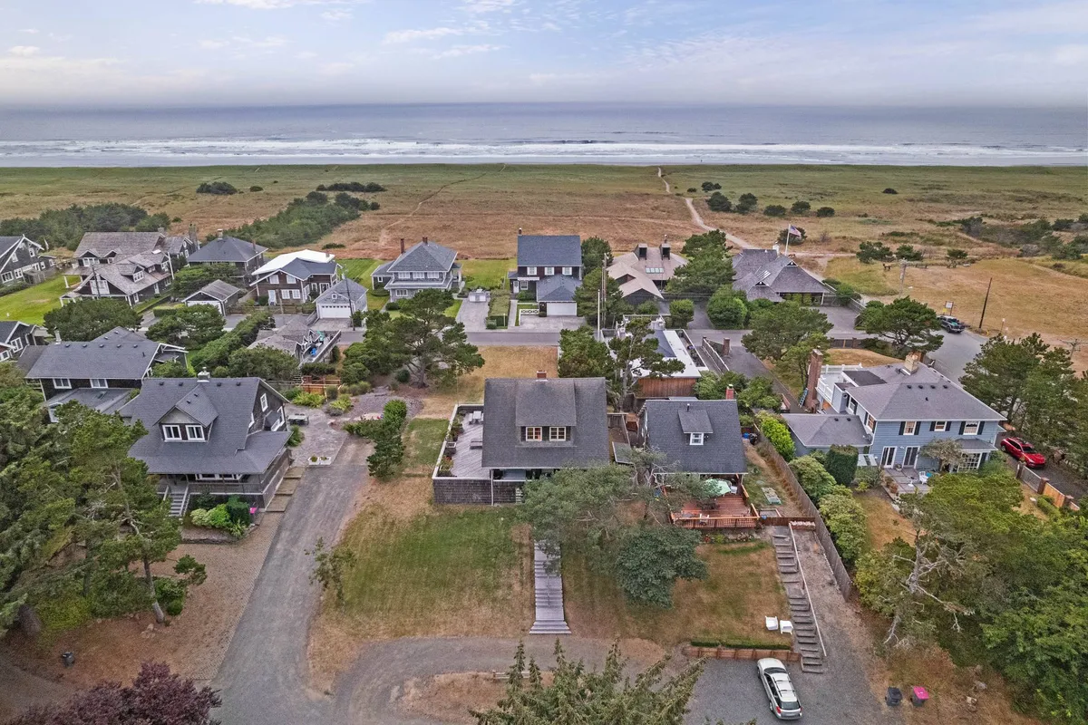 A serene beachfront scene with soft sands stretching out toward the ocean, framed by lush greenery and neighboring houses in the background. The scene invites the viewer to imagine family strolls, sandcastle building, and paddling in the gentle waves.