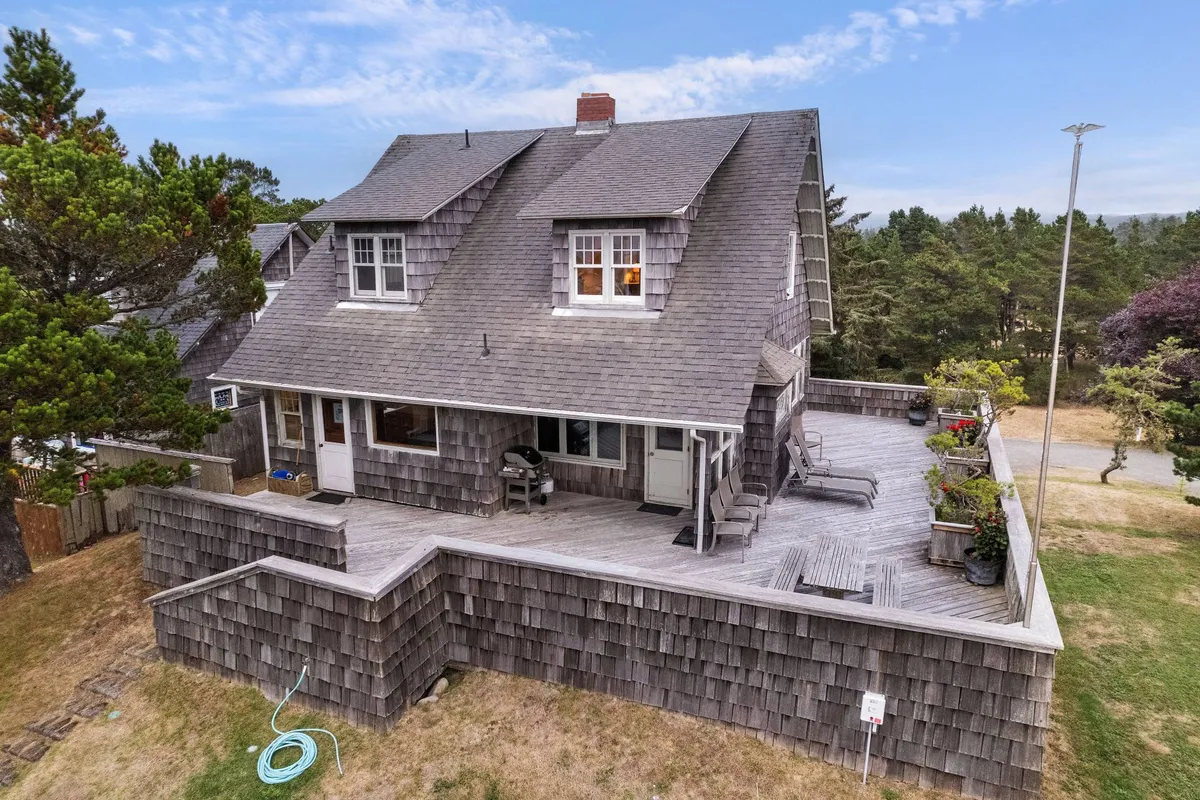 A welcoming wooden deck with outdoor seating, surrounded by greenery. The shingled exterior of the house and nearby plants create a relaxed and inviting atmosphere, suggesting a comfortable spot to unwind after a day at the beach.