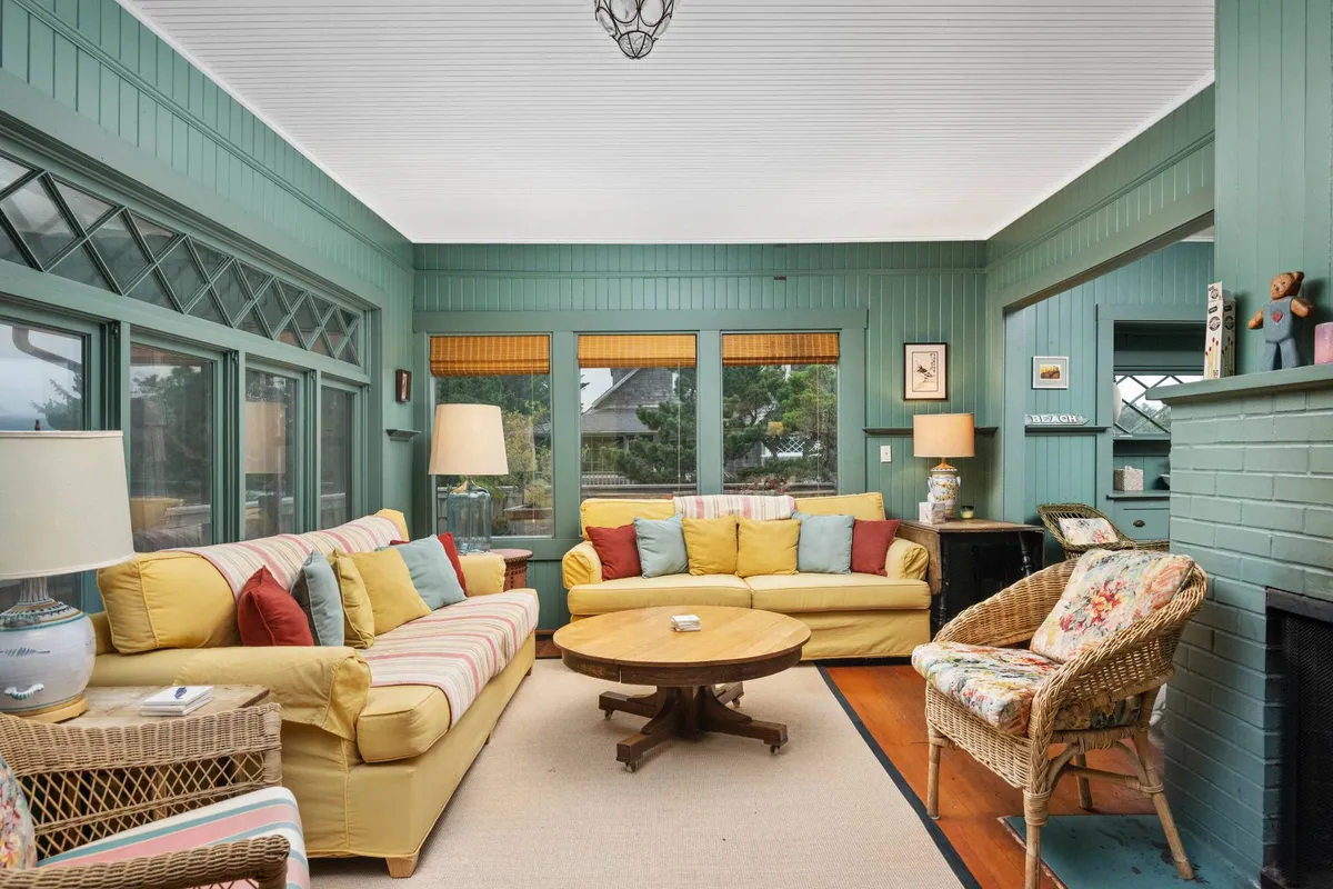 A sunlit living area with a yellow sofa, round coffee table, large windows flooding the space with natural light, a cozy wicker chair, and a table lamp. The room feels comfortable, perfect for relaxing with a book or gathering for board games.