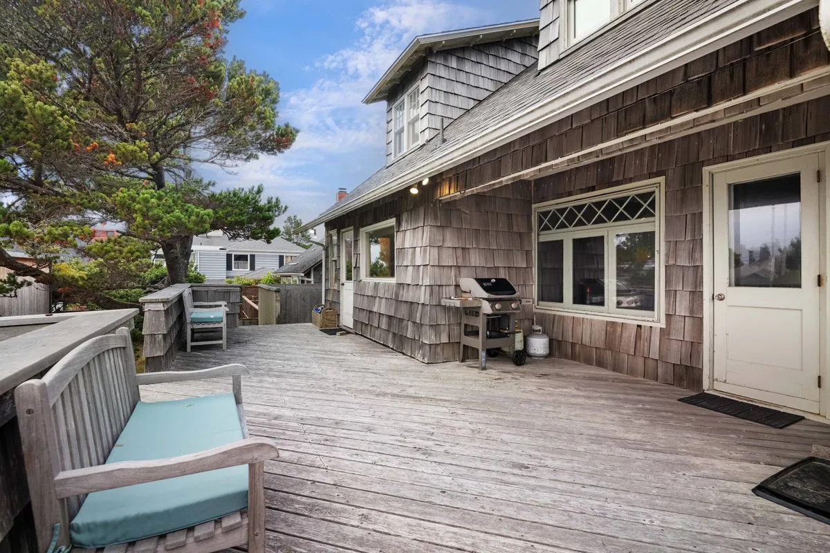 A welcoming wooden deck featuring a barbecue grill, wooden seating, and shingle siding. The deck area is bathed in natural light, suggesting a perfect spot for outdoor dining or lounging while taking in partial ocean views. Flower pots and greenery add a touch of color and life to the space, hinting at the possibilities for both lively gatherings and serene relaxation.